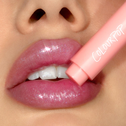 Shimmering pink lips wearing Cyberstar as a peach ColourPop stick applies the ColourPop So Juicy Plumping Gloss Balm with Peptides, showcasing glossy sparkle and a plumped, hydrated finish in a tight close-up.