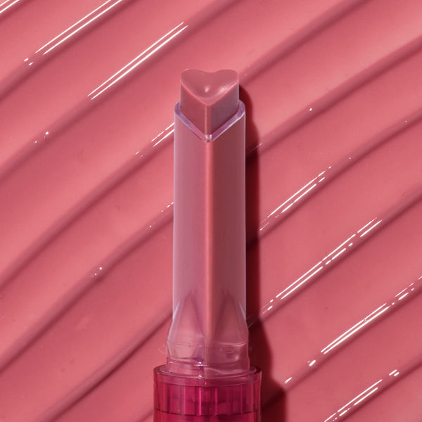 Heart-shaped tip of the Forbidden Love shade glides across a ribbed, glossy rose‑pink swatch, highlighting the shine and creamy payoff of the ColourPop So Juicy Plumping Gloss stick in a clear pink tube.