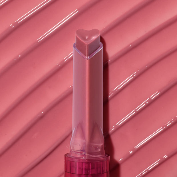 Heart-shaped tip of the Forbidden Love shade glides across a ribbed, glossy rose‑pink swatch, highlighting the shine and creamy payoff of the ColourPop So Juicy Plumping Gloss stick in a clear pink tube.
