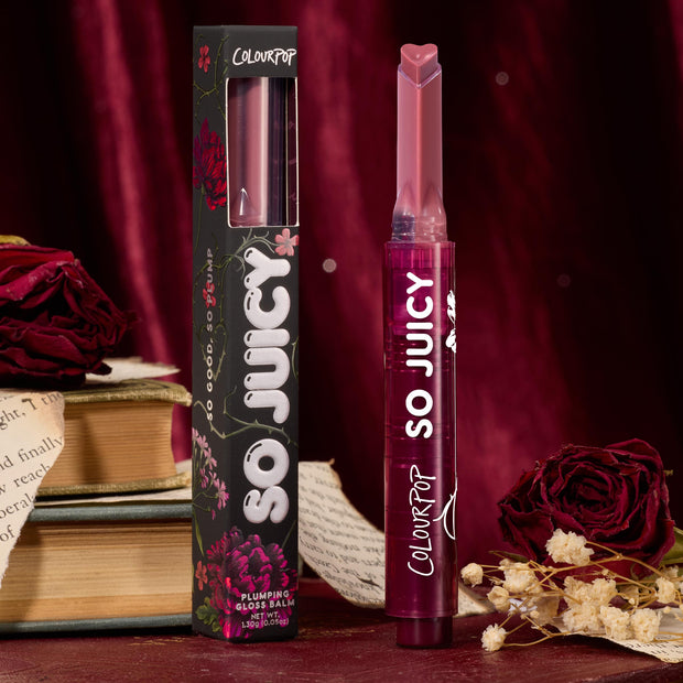 ColourPop So Juicy Plumping Gloss in Forbidden Love stands uncapped with a heart-shaped tip, shown beside its floral box on a burgundy velvet backdrop with dried roses and book pages, highlighting the deep berry gloss balm stick.