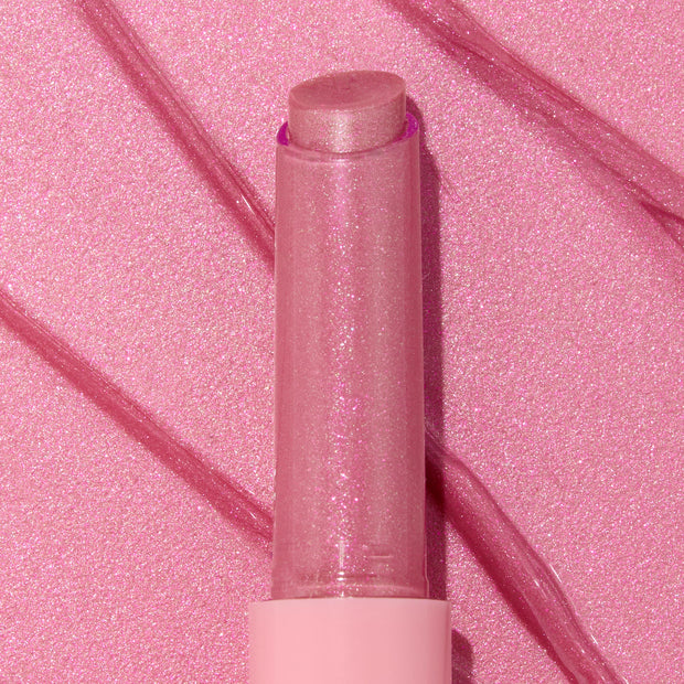 Shimmery pink Girly Pop stick of ColourPop So Juicy Plumping Gloss Balm with Peptides standing upright with the cap off, the glittery balm bullet catching light, set on a matching pink, glossy swatched background with diagonal streaks.