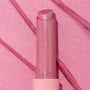 Shimmery pink Girly Pop stick of ColourPop So Juicy Plumping Gloss Balm with Peptides standing upright with the cap off, the glittery balm bullet catching light, set on a matching pink, glossy swatched background with diagonal streaks.