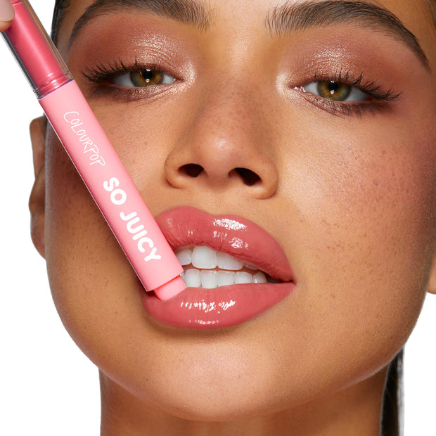 ColourPop So Juicy Plumping Gloss Balm with Peptides in Guava Glaze slicked onto full, glossy pink lips as the chubby pink tube is held to the mouth, close-up beauty shot with dewy skin and soft shimmer eye makeup.