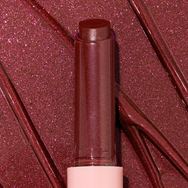 Gloss balm stick twisted up from a pink tube, L-O-V-E shade of the ColourPop So Juicy Plumping Gloss Balm with Peptides, shown close-up against glossy rose-plum shimmer swatches to highlight its dewy, plumping shine and sparkly finish.