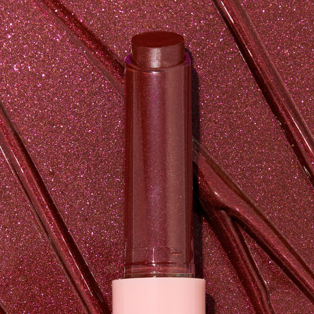 Gloss balm stick twisted up from a pink tube, L-O-V-E shade of the ColourPop So Juicy Plumping Gloss Balm with Peptides, shown close-up against glossy rose-plum shimmer swatches to highlight its dewy, plumping shine and sparkly finish.