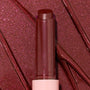 Gloss balm stick twisted up from a pink tube, L-O-V-E shade of the ColourPop So Juicy Plumping Gloss Balm with Peptides, shown close-up against glossy rose-plum shimmer swatches to highlight its dewy, plumping shine and sparkly finish.