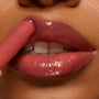 ColourPop So Juicy Plumping Gloss Balm with Peptides in PSL glides onto lips from a slim applicator, delivering a high-shine rosy brown finish and plumped look in a tight close-up of a mouth against warm skin.