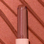 ColourPop So Juicy Plumping Gloss Balm with Peptides in shade Perfection standing upright with bullet extended, a rosy nude shimmer, in a clear cap and blush pink base, shown in macro against a matching metallic swatched background.
