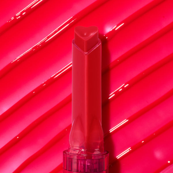 ColourPop So Juicy Plumping Gloss in Spice Level stands upright with a heart-shaped tip, showcasing a warm red shade against a glossy hot-pink swatch backdrop with diagonal ridges, emphasizing its high-shine, lip-plumping finish.