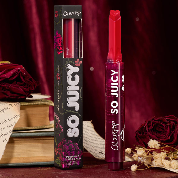 ColourPop So Juicy Plumping Gloss in Spice Level stands upright with a heart‑tipped stick, showing a deep berry red shade beside its black floral box, styled with vintage books and dried roses against a burgundy velvet backdrop.