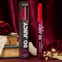 ColourPop So Juicy Plumping Gloss in Spice Level stands upright with a heart‑tipped stick, showing a deep berry red shade beside its black floral box, styled with vintage books and dried roses against a burgundy velvet backdrop.