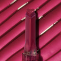 Heart-shaped tip Temptation shade lip gloss stick stands upright against a glossy ribbed hot‑pink swatch, showcasing rich pigment and shine from the ColourPop So Juicy Plumping Gloss.