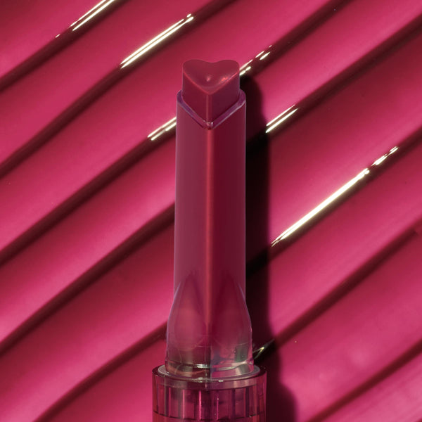 Heart-shaped tip Temptation shade lip gloss stick stands upright against a glossy ribbed hot‑pink swatch, showcasing rich pigment and shine from the ColourPop So Juicy Plumping Gloss.