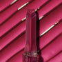 Heart-shaped tip Temptation shade lip gloss stick stands upright against a glossy ribbed hot‑pink swatch, showcasing rich pigment and shine from the ColourPop So Juicy Plumping Gloss.