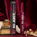 ColourPop So Juicy Plumping Gloss in Temptation stands upright with a heart-shaped tip beside its black floral box, a deep berry tube set among red velvet, roses, dried blooms, and vintage books for a romantic product shot.