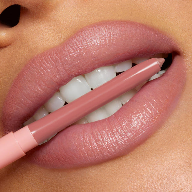 ColourPop So Juicy Plumping Lip Liner with Peptides in Love That lines plush, glossy lips, the rosy‑nude pencil held between teeth to showcase its precise tip and creamy payoff in a clean studio close‑up.