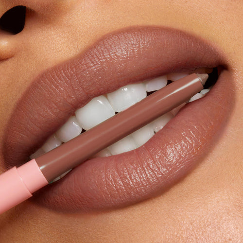 ColourPop So Juicy Plumping Lip Liner with Peptides in Tease defining full lips as the nude-brown pencil, with a blush-pink cap, is held between teeth, showcasing precise, creamy payoff and smooth contour in a studio close-up.