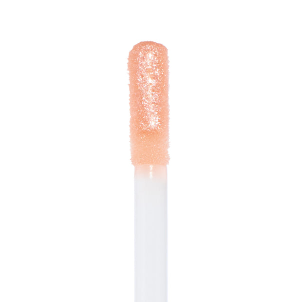 Doe-foot applicator coated in Sugar Cookie ColourPop Ultra Glossy Lip, a sheer peach-nude gloss with fine gold sparkle, glistening in macro close-up against a white backdrop.