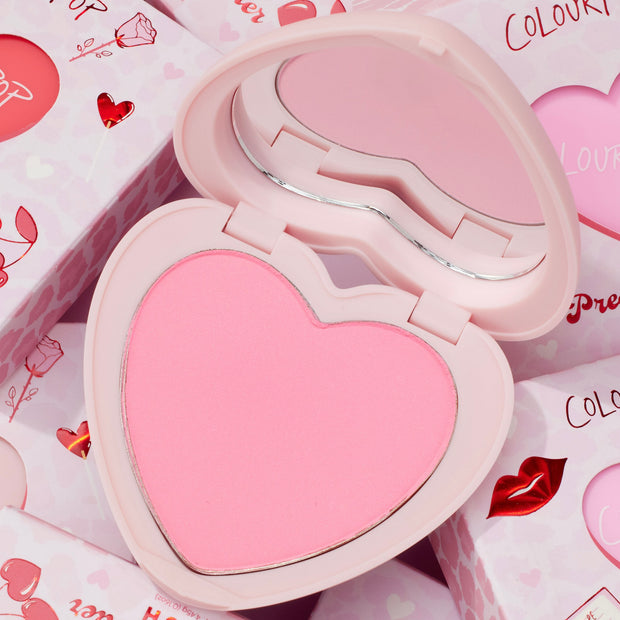 Sweetheart heart-shaped compact opens to reveal a soft baby pink pan and curved mirror, ColourPop Pressed Powder Blush, resting on Valentine-themed packaging with hearts, roses, and lip prints.