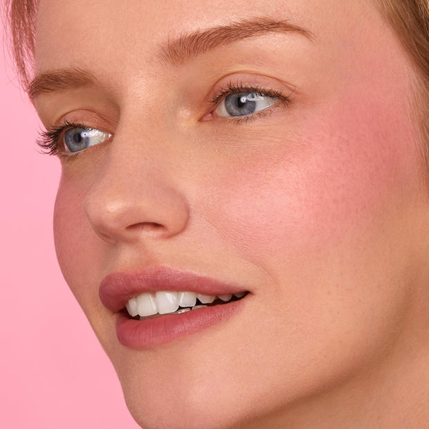 Rosy cheek and soft pink lips glow, showcasing the That’s So Rosy ColourPop Cheek & Lip Set on a close-up model; creamy blush and matching lip tint blend seamlessly with a dewy finish against a pastel pink background.