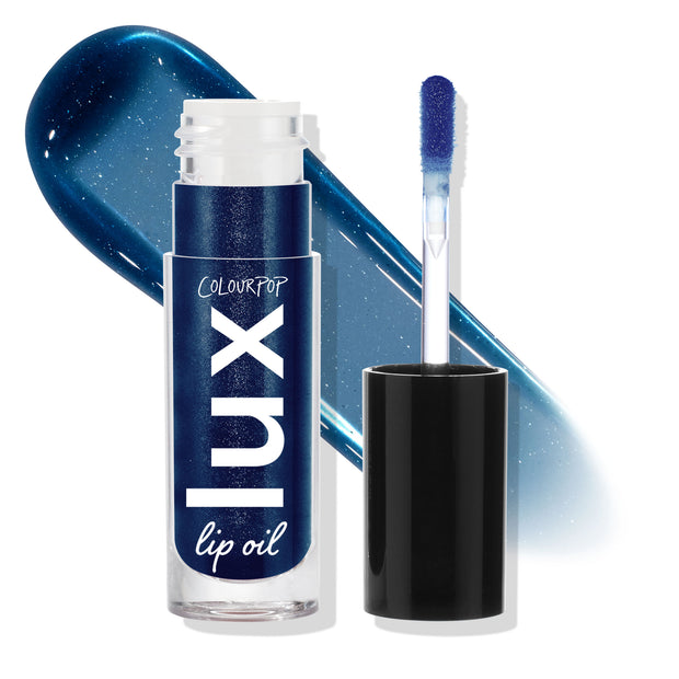 Team Edward ColourPop Lux Lip Oil standing with cap off and doe-foot applicator, set against a glossy midnight-blue shimmer swatch; clear tube labeled lux lip oil on a white studio background.