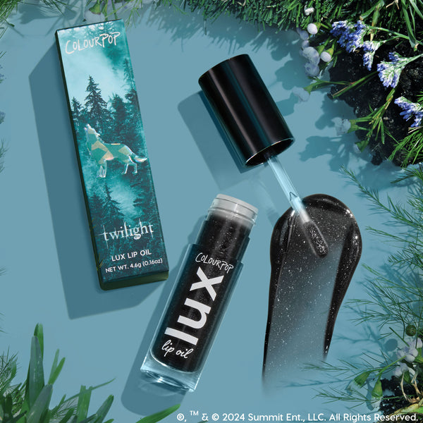 Team Jacob ColourPop Lux Lip Oil shown open with doe-foot applicator and a glossy black, silver-sparkle swatch, beside the Twilight-themed box with wolf and forest art, 4.6g (0.16oz), on a teal flat lay with pine and wildflower accents.