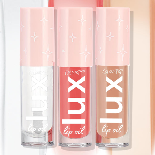 Three ColourPop Lux Lip Oil tubes stand side by side in the Top Hits bundle, displaying clear, coral pink, and warm nude shades with pink star-print caps against a soft pastel backdrop.