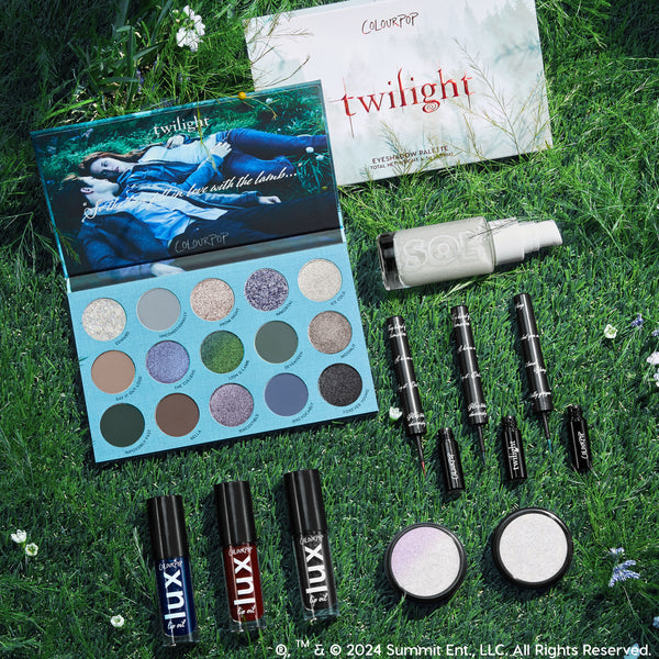 Twilight x ColourPop Full Collection arranged in a flatlay on grass with wildflowers, showcasing an open 16-pan cool-toned eyeshadow palette, black liquid liners, LUX lip oils, two iridescent highlighters, and a SOL body shimmer - ColourPop Full Collection Set.