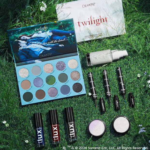 Twilight x ColourPop Full Collection arranged in a flatlay on grass with wildflowers, showcasing an open 16-pan cool-toned eyeshadow palette, black liquid liners, LUX lip oils, two iridescent highlighters, and a SOL body shimmer - ColourPop Full Collection Set.