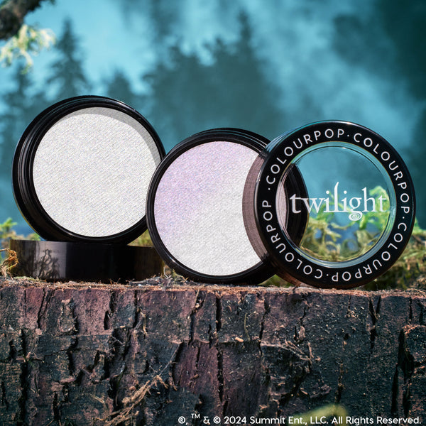 Meadow ColourPop Super Shock Highlighter compact opens to reveal an iridescent silver‑lilac pan, with a Twilight‑branded lid propped beside it, styled on a mossy tree stump against a misty forest backdrop.