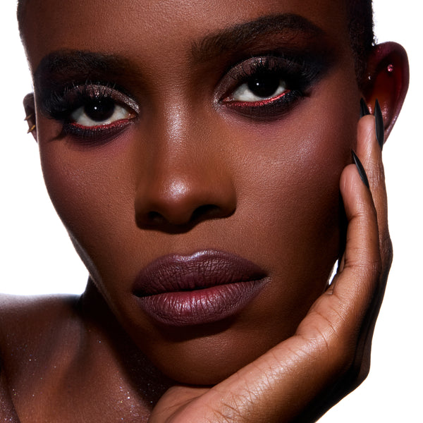 Model rests hand on face wearing deep plum lipstick, crimson-lined smokey eyeshadow, and glowing skin created with The Twilight Saga Eclipse X ColourPop full collection set, showcasing dramatic Twilight inspired glam beauty look on white background.