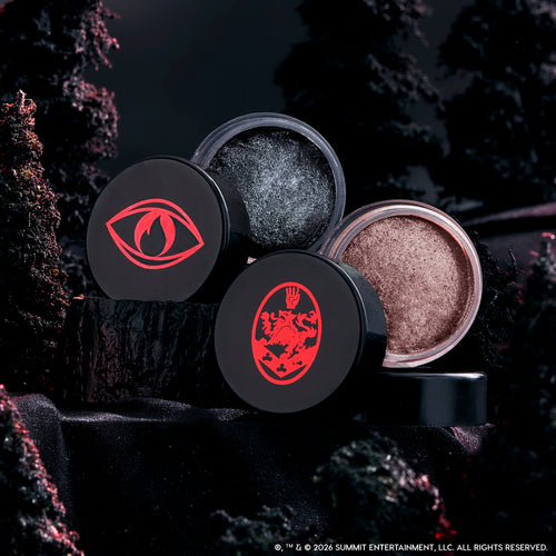 Coven Clash Jelly Much Duo eyeshadows sit open on black fabric, revealing shimmery charcoal and champagne shades in round pots with bold red Twilight-inspired symbols, styled against a dark, moody forest backdrop.