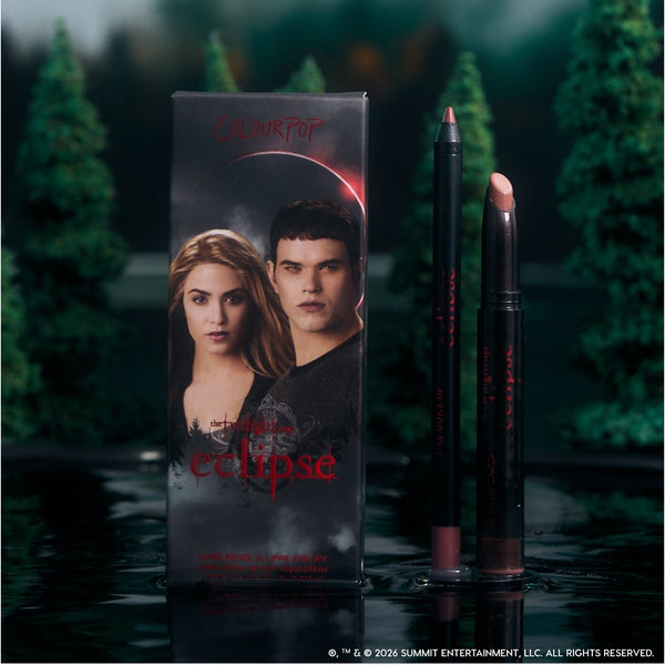 ColourPop Opposites Attract Lippie Stix and lip pencil from the Twilight Eclipse collection stand beside their character-adorned box on a reflective lake surface, surrounded by dark evergreen trees for a moody, cinematic makeup display.