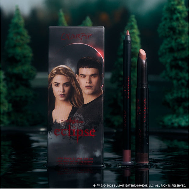 ColourPop Opposites Attract Lippie Stix and lip pencil from the Twilight Eclipse collection stand beside their character-adorned box on a reflective lake surface, surrounded by dark evergreen trees for a moody, cinematic makeup display.