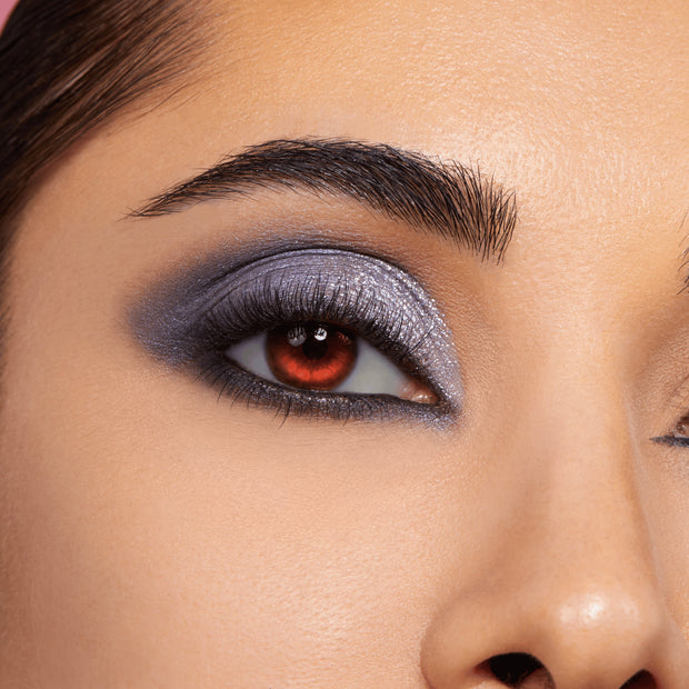 Smoky silver-lavender shimmer eyeshadow with charcoal blend and glitter accents covers a model’s lid, paired with bold lashes and a red contact lens, showcasing the Twilight Shadow Palette, a ColourPop Shadow Palette.