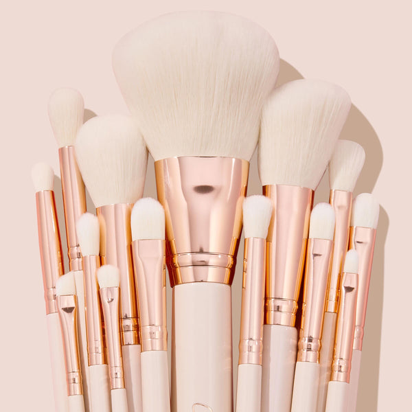 Ultimate Brush Roll, a ColourPop Makeup Brush Kit, fanned upright with soft white bristles, rose-gold ferrules, and cream handles, showcasing face and eye brushes for powder, blush, and eyeshadow blending on a blush-pink studio backdrop.
