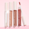 ColourPop Ultra Glossy Lip 20 Carats with doe‑foot applicator lifted, showing rosy nude shine, stands beside three closed ColourPop Ultra Glossy Lip tubes - clear, peach nude, and bronze shimmer - with star‑embossed caps on a soft pink backdrop.