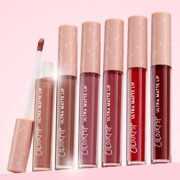 ColourPop Ultra Matte Lip Flurries shows the doe-foot wand lifted from the tube, revealing a nude-mauve liquid lipstick beside five companion Ultra Matte Lip shades from beige to deep red, with blush star-print caps on a soft pink backdrop.