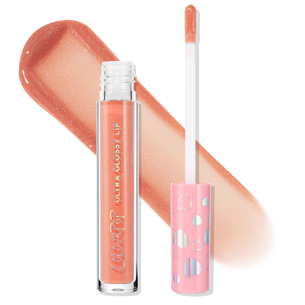 ColourPop Ultra Glossy Lip One Kiss lip gloss stands uncapped with a doe-foot applicator and a wide peachy-coral shimmer swatch behind, featuring a clear tube and pink cap with holographic hearts for a high-shine, sparkly finish.