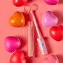 ColourPop Ultra Glossy Lip in One Kiss, a peachy‑pink shimmer, shown with open tube and applicator creating a heart-shaped swipe; pink cap with iridescent hearts; surrounded by red, pink, and orange heart props on a coral background.