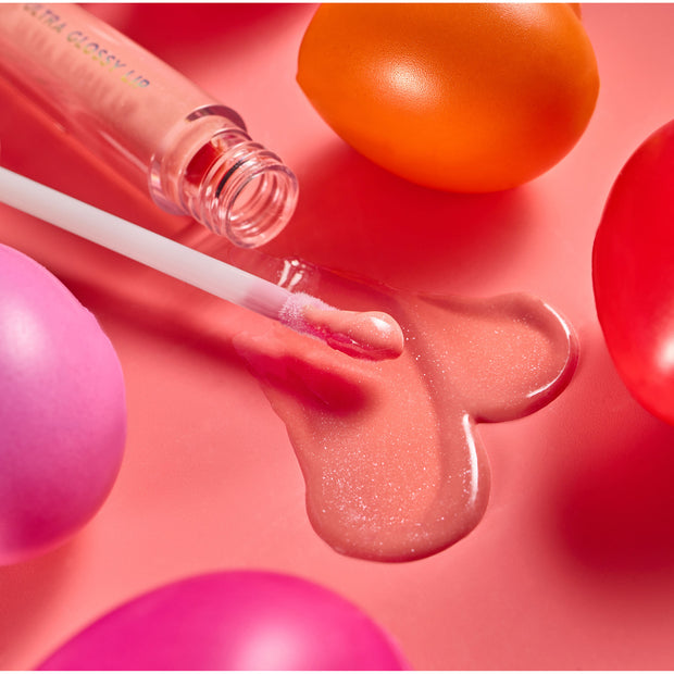 One Kiss Ultra Glossy Lip applicator swipes peachy‑pink shimmer gloss into a heart-shaped puddle beside its open tube on a coral surface with colorful rounded props; ColourPop Ultra Glossy Lip shows a high-shine, cushiony finish.