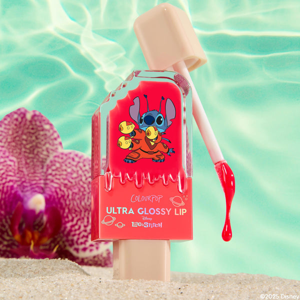 ColourPop Ultra Glossy Lip in Experiment 626 drips from the applicator wand onto beach sand, the clear rectangular bottle revealing bright coral gloss with a Stitch graphic, set poolside with turquoise water and a purple orchid.