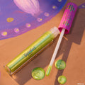 Pascal ColourPop Ultra Glossy Lip lying open with a doe-foot applicator and swirls of lime-green, gold-shimmer gloss, set on a Tangled-inspired painted backdrop, clear tube showing iridescent sheen and pink cap decorated with chameleon icons.