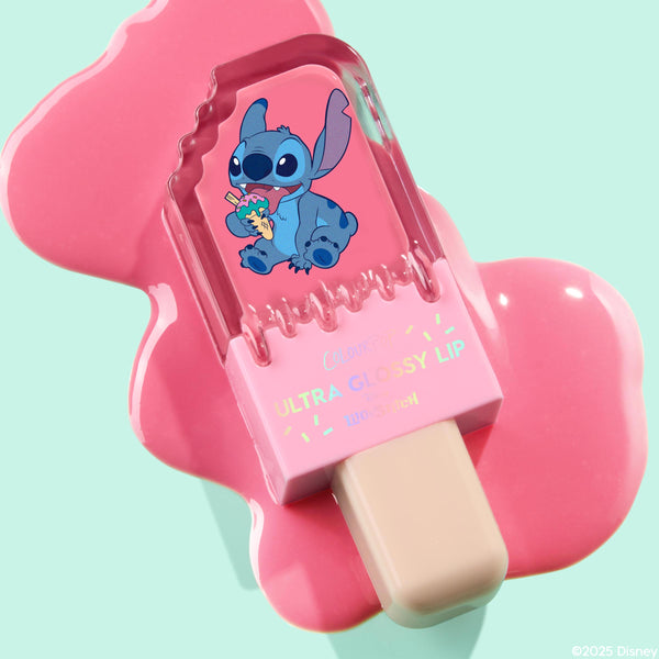 ColourPop Ultra Glossy Lip Super Sweet tube featuring Stitch rests on a melted pink gloss puddle, styled like a popsicle with a beige stick against a mint background, showcasing playful packaging and a bright candy-pink finish.