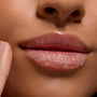 Glossy lips buffed with Neapolitan Sundae ColourPop Lip Scrub, sugar crystals exfoliating the lower lip as a fingertip applies the treatment in a close-up, revealing a smooth, hydrated finish.