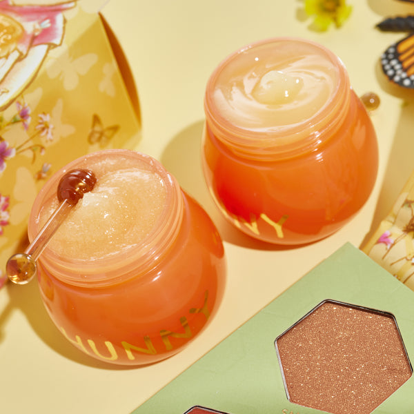 Sweet as Hunny Eye & Lip Set honey-pot orange jars sit open, revealing a sugar lip scrub and glossy balm with a mini honey dipper, beside a green shimmery eyeshadow palette and floral butterfly packaging on a pastel yellow surface; ColourPop Lip Care Kit.
