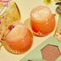 Sweet as Hunny Eye & Lip Set honey-pot orange jars sit open, revealing a sugar lip scrub and glossy balm with a mini honey dipper, beside a green shimmery eyeshadow palette and floral butterfly packaging on a pastel yellow surface; ColourPop Lip Care Kit.