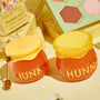 Hunny Pot lip care kit by ColourPop - two honey jar–shaped pots with yellow lids and HUNNY lettering - sit on a pale yellow surface beside a mini honey dipper, floral box, and makeup palette, showcasing the lip scrub and mask duo.