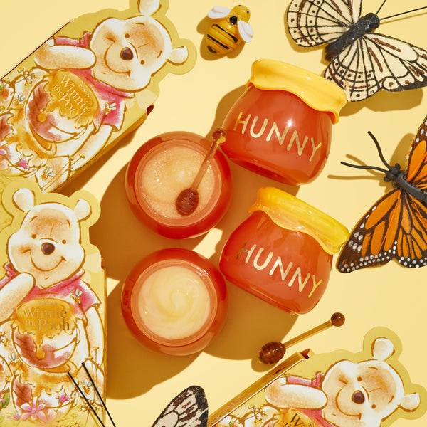 Sweet as Hunny ColourPop Eye & Lip Set displayed in a flat lay, featuring honey pot-shaped jars labeled HUNNY, two open pots of shimmery balm with mini honey-dipper applicators, and Winnie the Pooh packaging, butterflies, and a bee on a yellow background.