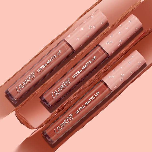Three ColourPop Ultra Matte Lip tubes in the Warmed Up bundle rest diagonally atop wide nude‑brown swatches, showcasing a full‑coverage ultra matte finish with clear barrels and blush pink star caps on a peach backdrop.